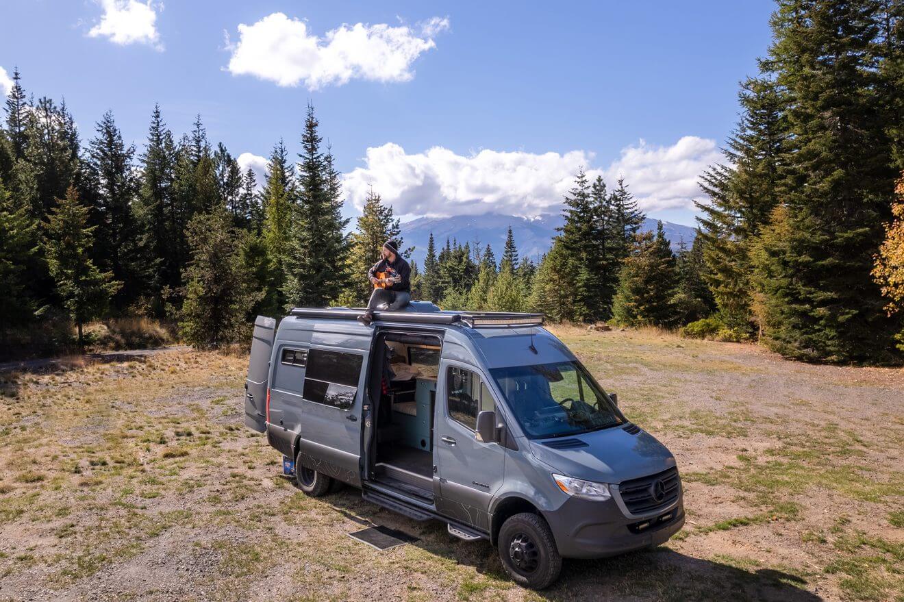 The exterior of a van in a field with a man sitting on the roof playing guitar