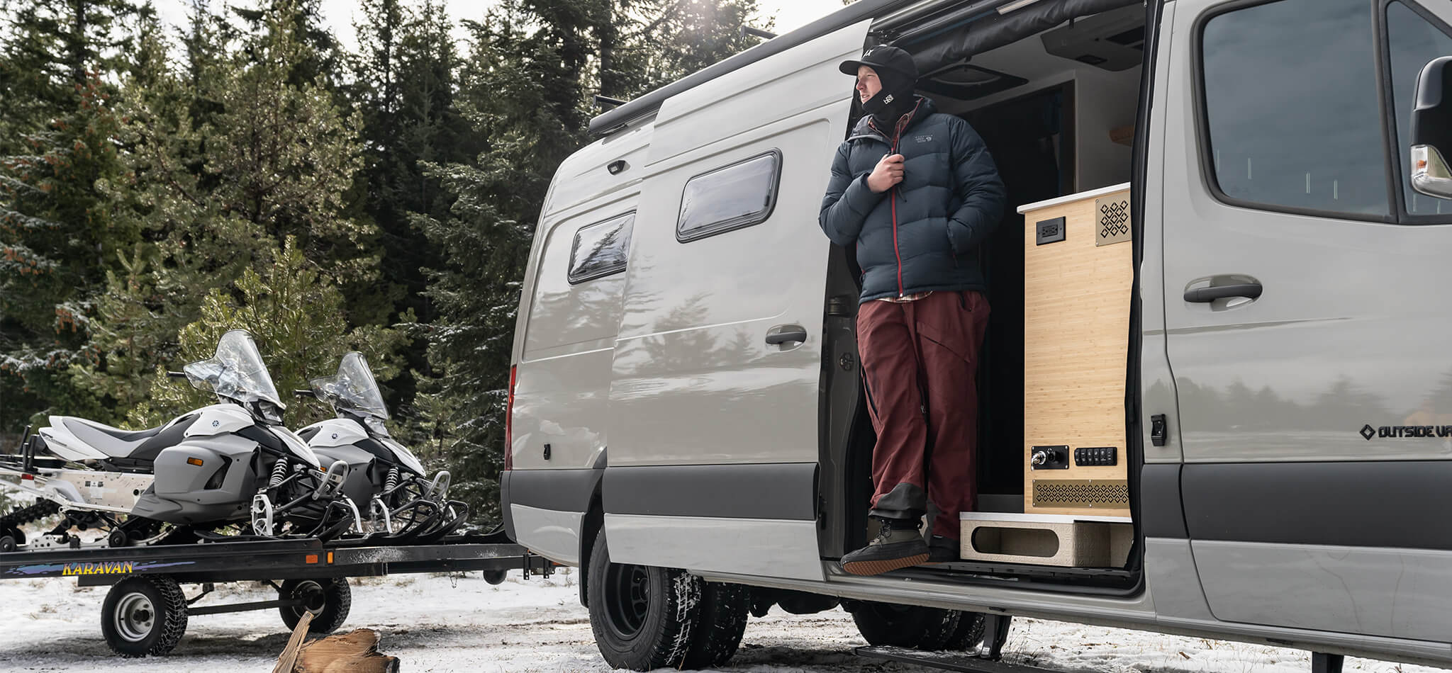 A man dressed in winter gear stands on the edge of his customized Sprinter camper van looking over a snow-covered landsape. There are two snowmobiles on a trailer in the background.