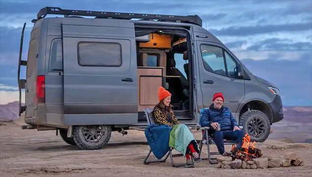 A couple enjoy a camp fire in desert scene sitting next to Winnebago Revel van.